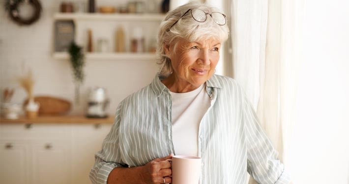 older lady stands with coffee in bright apartment